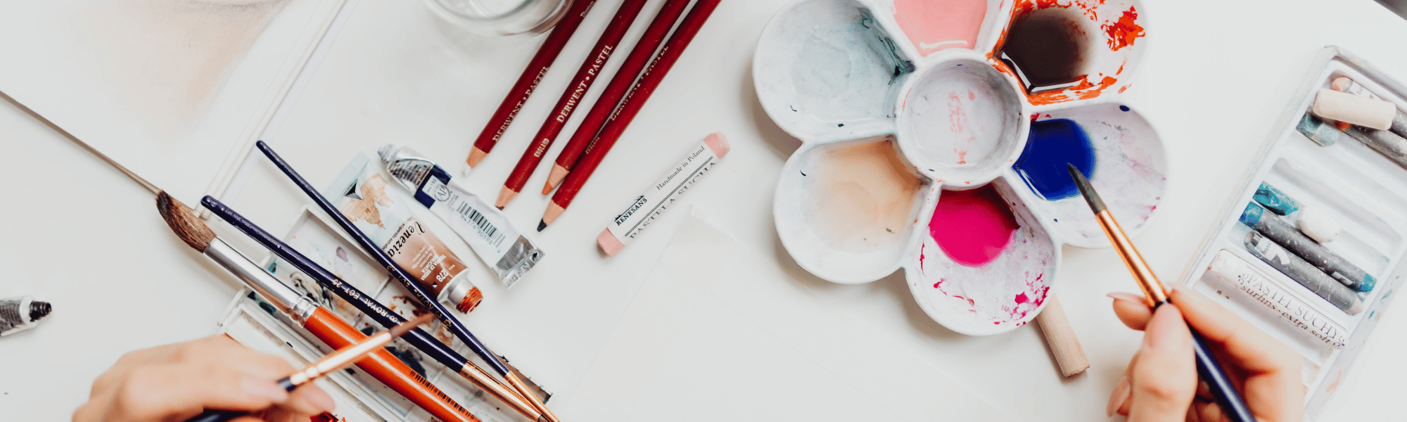 Artpack creates adult craft kits for DIY craft enthusiasts. Image shows two hands with craft tools and an assortment of craft supplies and materials spread out on the table.